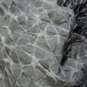 May include: Close-up of a textured gray fabric with a quilted pattern. The fabric has a three-dimensional effect, with raised sections and a network of lighter-colored lines creating a grid-like design. The material appears soft and pliable.
