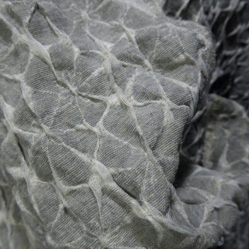 May include: Close-up of a textured gray fabric with a quilted pattern. The fabric has a three-dimensional effect, with raised sections and a network of lighter-colored lines creating a grid-like design. The material appears soft and pliable.