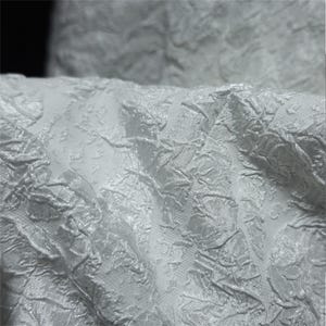 Puede incluir: Primer plano de una tela texturizada de color blanco roto. La tela tiene un patrón arrugado en relieve, creando una textura sutil e irregular. El material parece suave y se drapea suavemente, lo que sugiere que podría usarse para ropa o decoración del hogar.