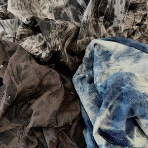 Könnte beinhalten: Nahaufnahme von mehreren Stoffmustern in Grau-, Braun- und Blautönen. Die Stoffe haben einen Batik- oder Marmoreffekt mit dezenten silbernen Fadenakzenten. Die Textilien sind gebündelt und gefaltet, wodurch ihre Textur zur Geltung kommt.