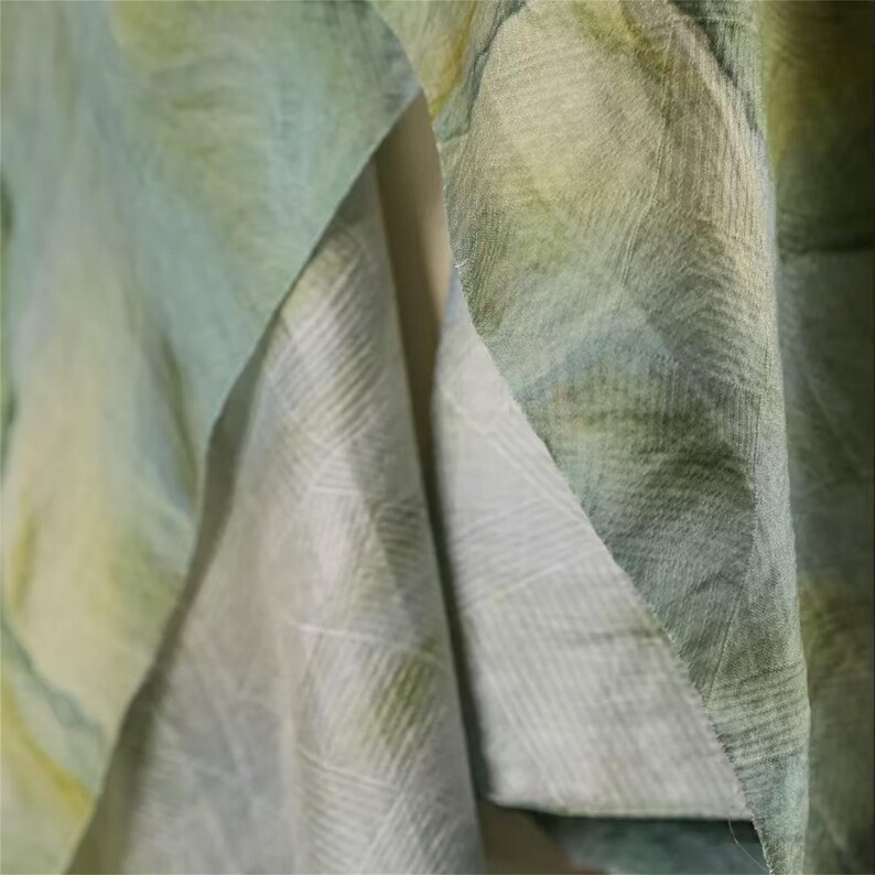 May include: Close-up of layered fabric in shades of green, blue, and yellow. The fabric has a subtle, textured pattern, with visible weave details. The folds and layers create depth and visual interest, suggesting a soft, flowing material.