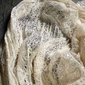 May include: An off-white, semi-sheer fabric featuring an intricate, open-knit mesh pattern. The material is embellished with small, reflective sequins scattered throughout the textured surface, creating a delicate, layered, and dimensional appearance.