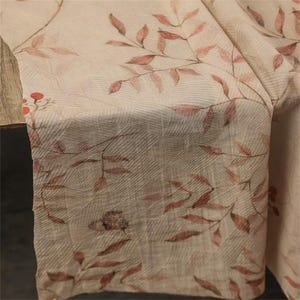 Op de afbeelding: Een beige stof met een delicaat patroon van rode en bruine bladeren en takken. De stof heeft een textuur, met een subtiel rasterachtig ontwerp. De bladeren zijn gedetailleerd en cre&euml;ren een natuurlijk, botanisch thema.