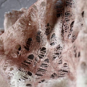 Puede incluir: Primer plano de una tela delicada de tejido abierto en tonos marrón claro y beige. La tela tiene un aspecto texturizado, casi de encaje, con numerosos agujeros irregulares, creando un efecto transparente. El material parece suave y ligero.