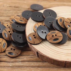 May include: A collection of wooden buttons in various shades of brown and black. Some buttons are solid black with two holes, while others are light brown with carved designs. The buttons are scattered on a wooden surface.