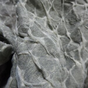 May include: Close-up of a textured, gray fabric with a pattern of raised, interconnected shapes. The fabric has a soft, almost velvety appearance, with subtle variations in tone and texture. The design creates a sense of depth and visual interest.