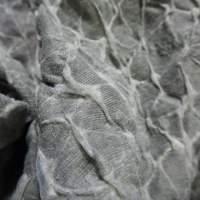 May include: Close-up of a textured, gray fabric with a pattern of raised, interconnected shapes. The fabric has a soft, almost velvety appearance, with subtle variations in tone and texture. The design creates a sense of depth and visual interest.