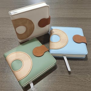 May include: Three square notebooks in white, light blue, and green. Each notebook features a large, stylized letter "D" design in a contrasting color, a brown leather closure, and a ribbon bookmark. The notebooks are displayed on a wooden surface.