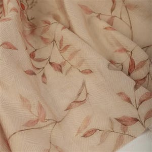 Op de afbeelding: Close-up van een beige stof met een delicaat bloemenpatroon. Het ontwerp toont takken in aquarel-stijl met roze en bruine bladeren. De stof heeft een subtiele, getextureerde weefsel, waardoor een zachte en luchtige uitstraling ontstaat.