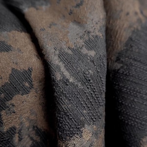 May include: Close-up of a textured fabric with a pattern in shades of brown and black. The fabric appears to be folded, revealing the intricate weave and subtle variations in colour and texture. The design has a mottled appearance.