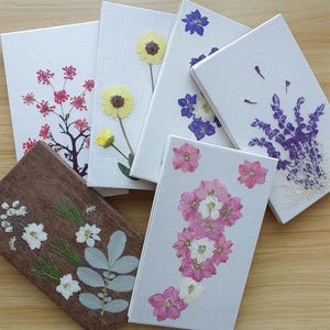 May include: A collection of handmade notebooks with pressed flowers. The covers are white and brown, adorned with various dried flowers in shades of pink, yellow, purple, and white. The notebooks are arranged on a wooden surface.