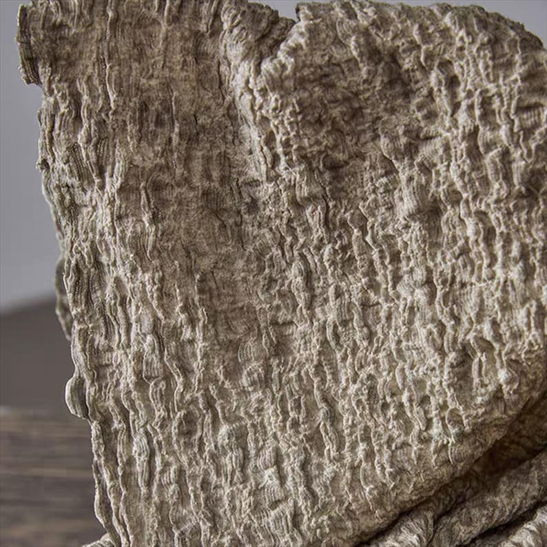 May include: Close-up of a textured, beige fabric with a crinkled appearance. The material has a rough, organic look, with visible ridges and folds. The fabric's surface is uneven, creating shadows and highlights.