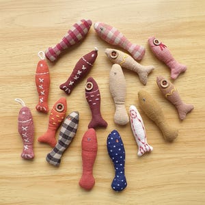 May include: A collection of handmade fabric fish in various colors and patterns. The fish feature details like button eyes, embroidered accents, and different fabric textures, including plaid and polka dots. Some have small loops for hanging.