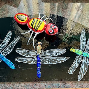 May include: Three glass insect ornaments, a red and yellow bee and two blue and green dragonflies, on a black background.