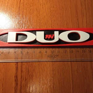 Puede incluir: Un letrero impreso en 3D de color rojo y blanco con la palabra "DUO" en letras blancas y "TM" en letras rojas. El letrero mide aproximadamente 45 cm de largo y est&aacute; sobre una superficie de madera.