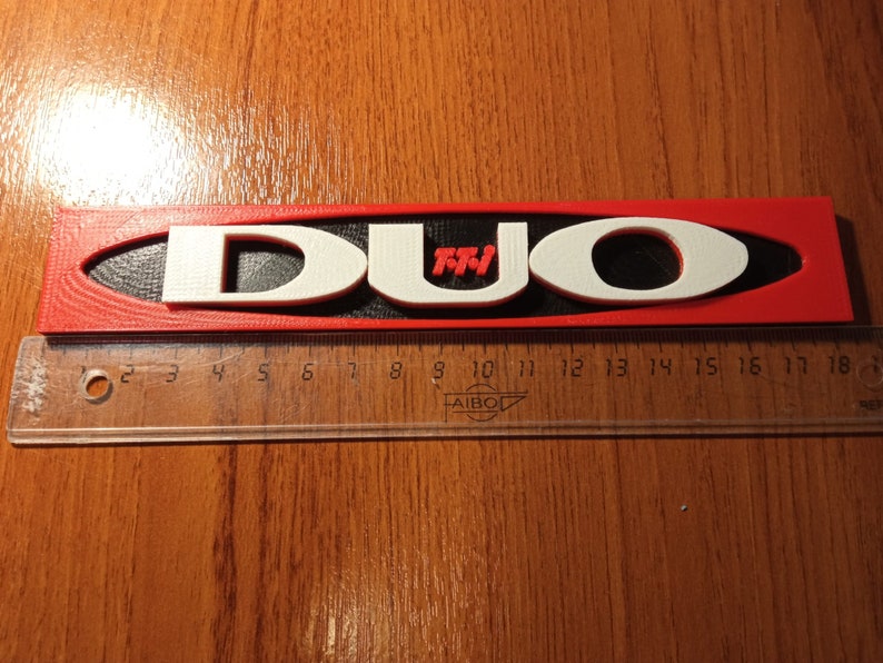 Puede incluir: Un letrero impreso en 3D de color rojo y blanco con la palabra "DUO" en letras blancas y "TM" en letras rojas. El letrero mide aproximadamente 45 cm de largo y est&aacute; sobre una superficie de madera.