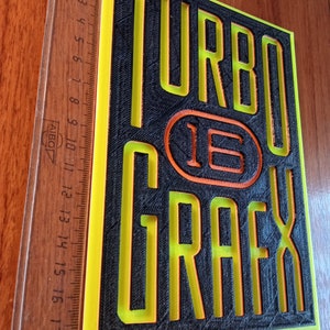 Puede incluir: Un letrero impreso en 3D en negro y amarillo con el texto "TURBO 16 GRAFX" en una fuente en negrita y de bloques.