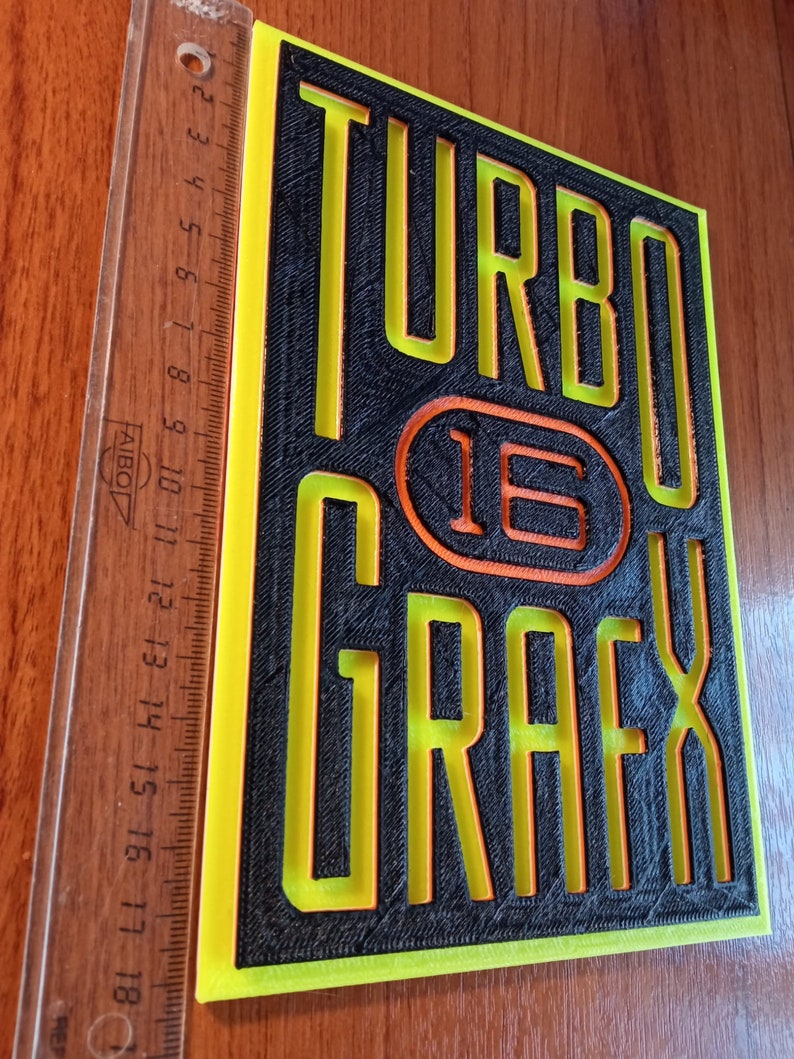Puede incluir: Un letrero impreso en 3D en negro y amarillo con el texto "TURBO 16 GRAFX" en una fuente en negrita y de bloques.