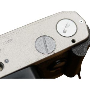 Puede incluir: Primer plano de la parte trasera de una cámara vintage. El cuerpo de la cámara es plateado con un acabado texturizado. Un detalle circular grabado está centrado en la parte trasera, y un pequeño dial circular blanco es visible. La palabra "MADE" está estampada en la cámara.