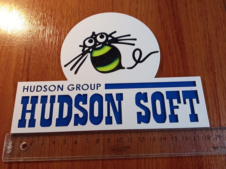 Puede incluir: Un cartel blanco con letras azules que dice "HUDSON GROUP" y "HUDSON SOFT". Una abeja de dibujos animados con rayas negras y amarillas y ojos grandes est&aacute; por encima del texto.