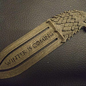 May include: A metallic gold bookmark with the text "WINTER IS COMING" engraved on it. The bookmark features a wolf head design at the top. The bookmark is made of a metallic material and is designed to be inserted into a book.