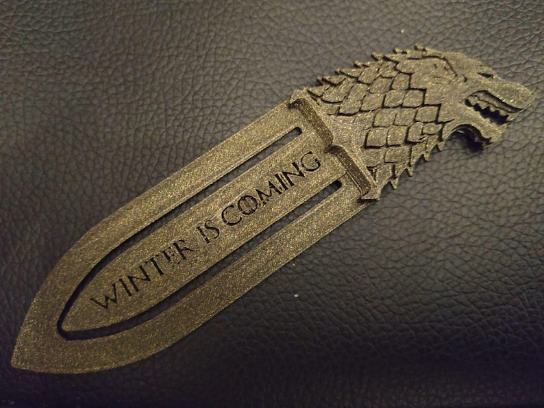May include: A metallic gold bookmark with the text "WINTER IS COMING" engraved on it. The bookmark features a wolf head design at the top. The bookmark is made of a metallic material and is designed to be inserted into a book.