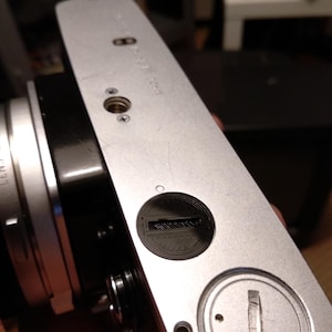 Puede incluir: Primer plano de un cuerpo de cámara plateado vintage. La cámara presenta una palanca de avance de película negra, un dial circular negro y una base plateada con varios tornillos y marcas. La lente es visible en el lado izquierdo.