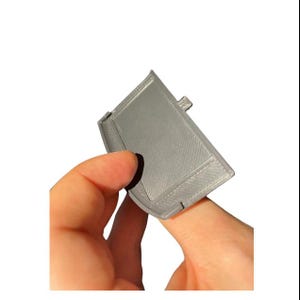 Può includere: Un oggetto rettangolare grigio stampato in 3D con un bordo curvo e una superficie strutturata. L'oggetto ha una piccola linguetta su un lato ed è tenuto da un dito. L'oggetto sembra essere una piccola custodia o supporto.