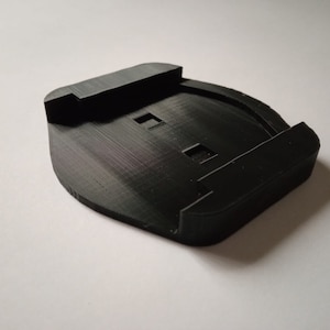 Puede incluir: Una base de plástico negro impresa en 3D con dos recortes rectangulares. La base está diseñada para ser utilizada como soporte para una cámara u otro dispositivo.