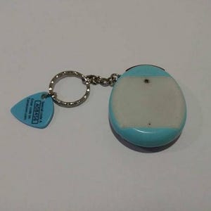 May include: A keychain with a light blue and white electronic device and a blue guitar pick-shaped tag. The tag has text and a silver key ring and chain. The device is oval-shaped.