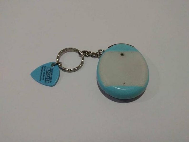 May include: A keychain with a light blue and white electronic device and a blue guitar pick-shaped tag. The tag has text and a silver key ring and chain. The device is oval-shaped.