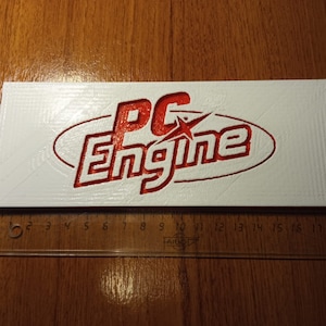Puede incluir: Un letrero rectangular blanco con un logotipo 3D rojo que dice "PC Engine" con una estrella en la "C".