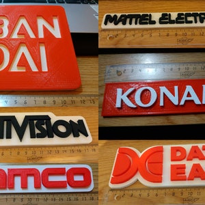 Puede incluir: Seis logotipos de empresas de videojuegos impresos en 3D: Bandai, Mattel Electronics, Konami, Activision, Namco y Data East. Los logotipos est&aacute;n impresos en rojo, blanco y negro sobre un fondo blanco.