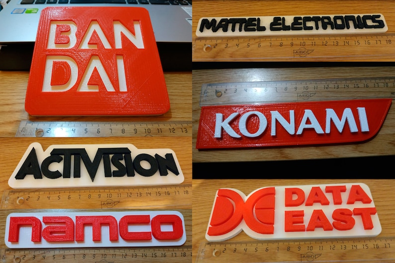 Puede incluir: Seis logotipos de empresas de videojuegos impresos en 3D: Bandai, Mattel Electronics, Konami, Activision, Namco y Data East. Los logotipos est&aacute;n impresos en rojo, blanco y negro sobre un fondo blanco.