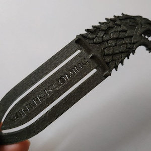May include: A dark grey bookmark with a wolf head design and the text "WINTER IS COMING". The bookmark is made of a solid material. A unique accessory for book lovers. The bookmark is approximately 15 cm in length.