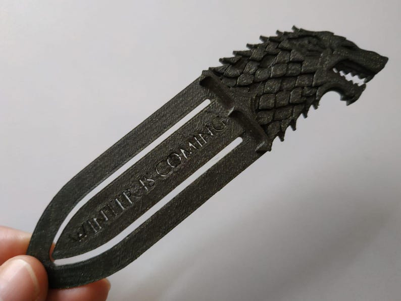 May include: A dark grey bookmark with a wolf head design and the text "WINTER IS COMING". The bookmark is made of a solid material. A unique accessory for book lovers. The bookmark is approximately 15 cm in length.