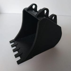 May include: A black 3D-printed excavator bucket, featuring a toothed edge and three holes at the top. The bucket has a curved design, possibly a toy or model for construction enthusiasts.