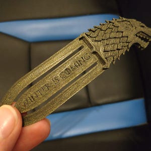 May include: A gold-coloured bookmark with the direwolf emblem from Game of Thrones. The words "WINTER IS COMING" are engraved on the bookmark. The bookmark is held by a hand.