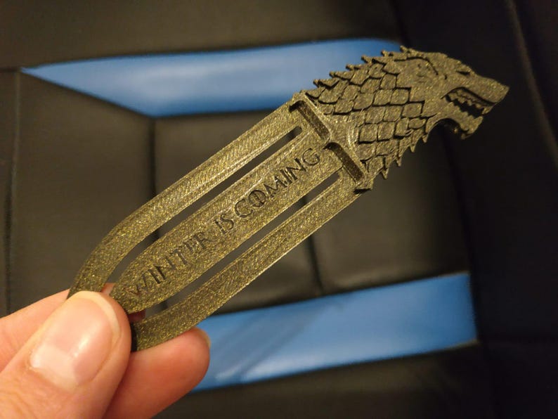 May include: A gold-coloured bookmark with the direwolf emblem from Game of Thrones. The words "WINTER IS COMING" are engraved on the bookmark. The bookmark is held by a hand.