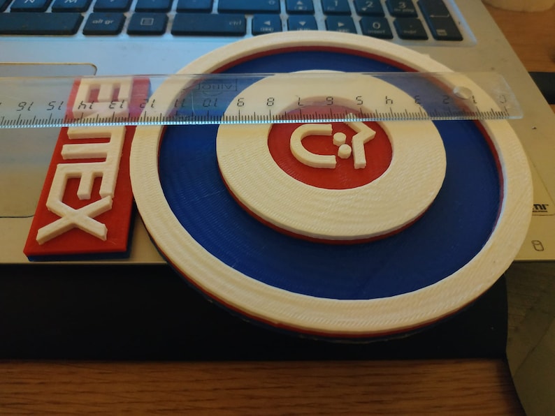 Puede incluir: Un logotipo impreso en 3D blanco, azul y rojo con el texto "Emtex" y una flecha estilizada que apunta hacia la derecha. El logotipo est&aacute; sobre una superficie de madera.