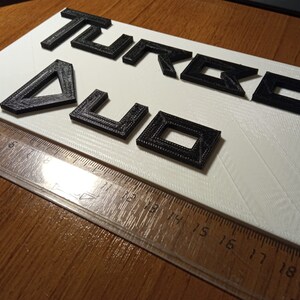 Puede incluir: Letras negras impresas en 3D que deletrean "TURBO 4U" sobre un fondo blanco.