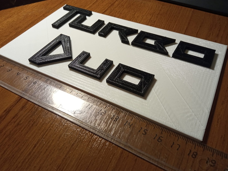 Puede incluir: Letras negras impresas en 3D que deletrean "TURBO 4U" sobre un fondo blanco.