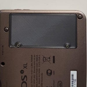 Puede incluir: Primer plano de la parte trasera de una consola Nintendo DS XL de color bronce. Una cubierta rectangular negra está asegurada con dos tornillos. La consola tiene texto, incluyendo "DS XL" y especificaciones técnicas.