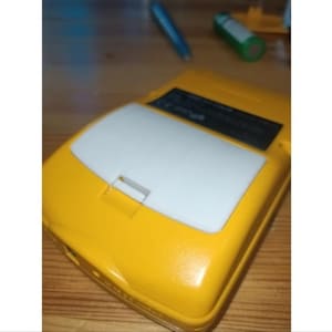 Puede incluir: Un dispositivo electrónico portátil amarillo con un panel rectangular blanco en la parte superior. El dispositivo tiene una pequeña abertura rectangular y una etiqueta negra. Un bolígrafo azul y una batería verde están en el fondo.