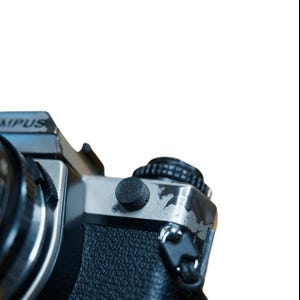 Puede incluir: Primer plano de una cámara Olympus vintage. El cuerpo de la cámara es negro y plateado, con un agarre texturizado. La parte superior presenta un dial negro y un botón plateado. La palabra "Olympus" es visible en la parte superior de la cámara.