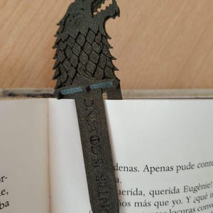 May include: A dark grey bookmark with a wolf's head design and the text "WINTER IS COMING." The bookmark is placed in a book, with the wolf's head showing above the pages. The bookmark is made of a textured material.