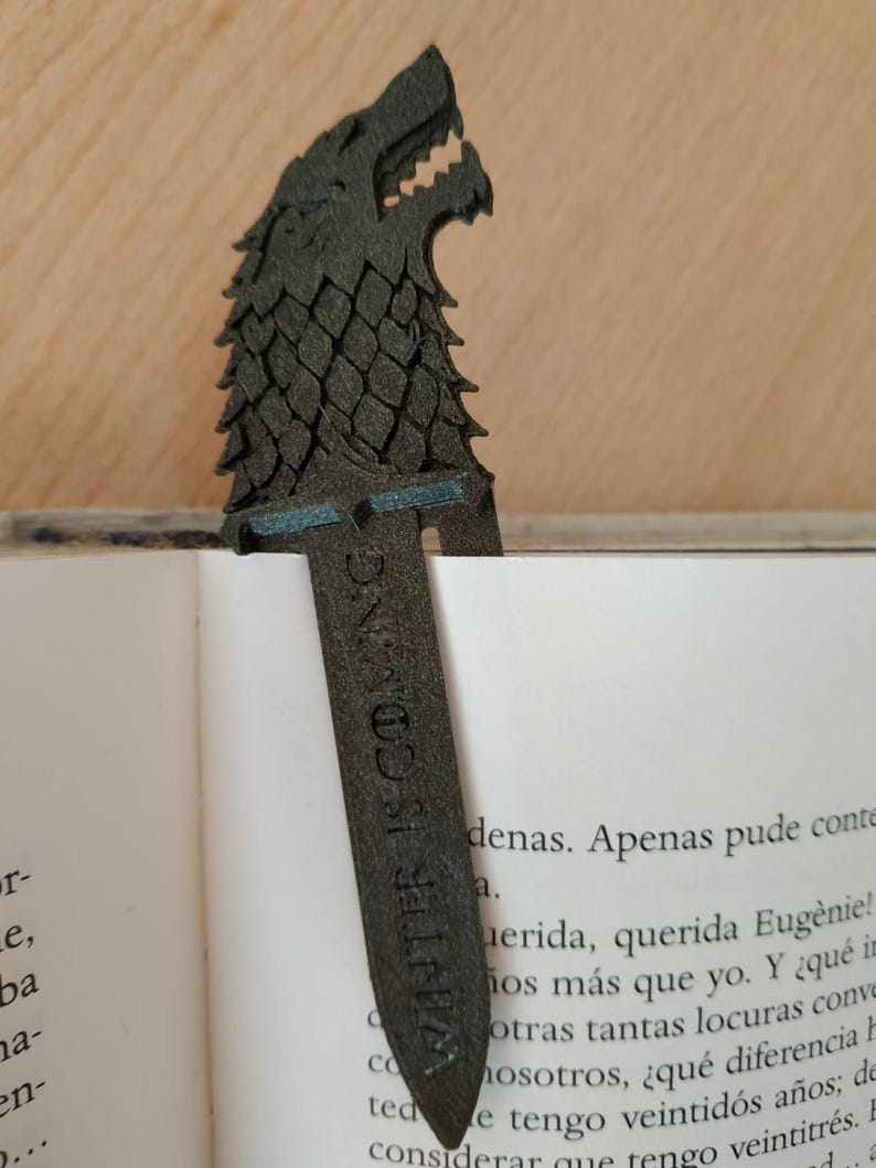 May include: A dark grey bookmark with a wolf's head design and the text "WINTER IS COMING." The bookmark is placed in a book, with the wolf's head showing above the pages. The bookmark is made of a textured material.