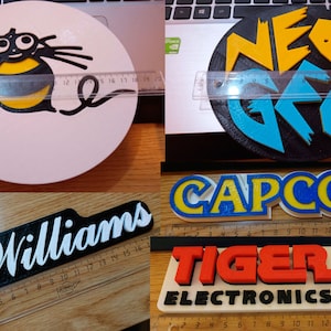 Puede incluir: Una colecci&oacute;n de logotipos impresos en 3D, incluyendo Williams, Capcom, Tiger Electronics y Neo Geo. Los logotipos est&aacute;n impresos en varios colores, incluyendo negro, blanco, rojo, azul y amarillo.