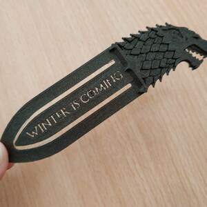 May include: A black bookmark featuring a wolf head design and the text "WINTER IS COMING". The bookmark is held in a hand. The bookmark is made of a dark material.