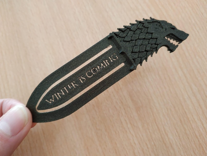 May include: A black bookmark featuring a wolf head design and the text "WINTER IS COMING". The bookmark is held in a hand. The bookmark is made of a dark material.